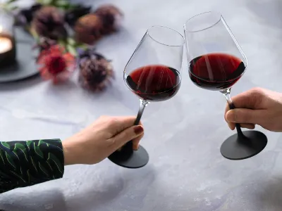 Deux personnes trinquent avec deux verres à vin rouge remplis Manufacture, avec un pied noir, de Villeroy & Boch.