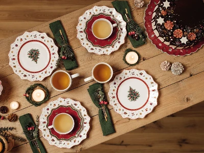 A wooden table with Christmas plates, green napkins, cups of tea, candles and a chocolate cake.
