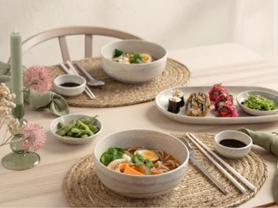 Una mesa puesta para dos personas con piezas Perlemor Una mesa puesta para dos personas con cuencos con ramen, rollitos de sushi, edamame, palillos y salsas para mojar de la colección Perlemor sobre manteles individuales tejidos.
