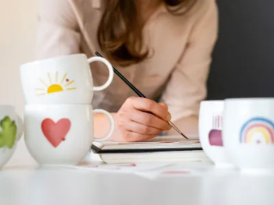  Des tasses With Love avec différents motifs, tandis qu’une femme peint en arrière-plan.