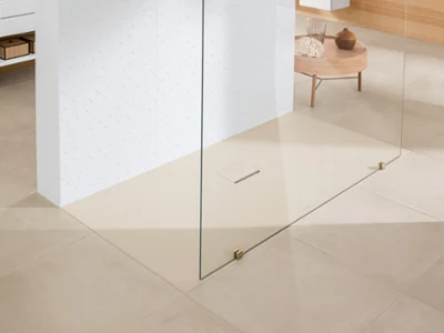 Une salle de bains minimaliste avec des carreaux de sol beiges, une douche à l’italienne séparée par une plaque en verre, un mur carrelé blanc et une petite table en bois avec des objets décoratifs en arrière-plan.