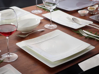 The Villeroy & Boch Modern Grace dining set consisting of square plates, wine glasses, bowls and cutlery arranged on a wooden surface with fern accents. A glass of red wine adds the finishing touch to the ambience.