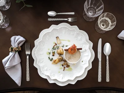 Des couverts de table Boston avec de la vaisselle de Noël Toy’s Delight Une table de salle à manger sophistiquée présente les assiettes « Boston » de Villeroy & Boch, artistiquement décorées de motifs ondulés, et complétées par des verres en cristallin, de couverts de table argentés richement décorés et de serviettes décoratives sur une table en bois foncé.