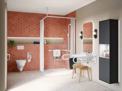 Une salle de bains accessible avec un carrelage orange hexagonal, une douche à l’italienne avec des poignées et un siège rabattable, un lavabo mural avec des rampes de sécurité, un miroir rétro-éclairé, des toilettes avec poignées et des unités de rangement.