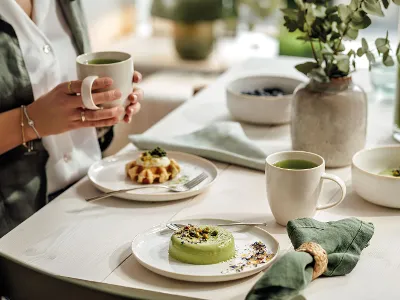 Une personne assise à une table de salle à manger tient une tasse à thé Crafted avec différents plats servis, dont un dessert vert, des gaufres et un bol de soupe.