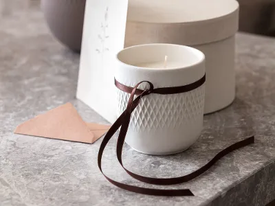 Une bougie parfumée Manufacture blanche de Villeroy & Boch emballée en cadeau avec un ruban marron et une boîte claire en arrière-plan.