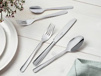 A neatly arranged stainless steel flatware set with knives, forks, and spoons on a light-colored wooden surface. Next to it are a white plate and a green napkin, with a flower arrangement in the top left-hand corner.