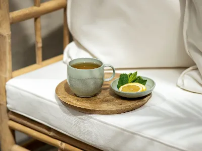 Une tasse de thé Perlemor est posée sur un plateau en bois, à côté d'un petit plat contenant une tranche de citron et des feuilles de menthe, sur une chaise blanche en bambou.
 