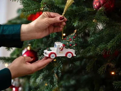 A person hangs a small truck-shaped ornament from Villeroy  and Boch on a decorated Christmas tree adorned with red baubles and lights.