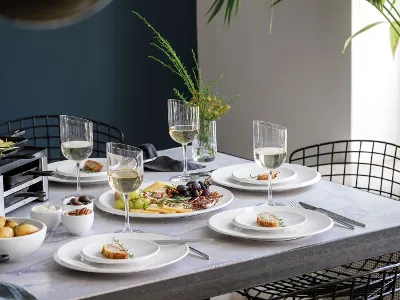 Un tavolo da pranzo con piatti bianchi NewMoon, bicchieri da vino, formaggio, frutta e antipasti, pronto per un pasto.