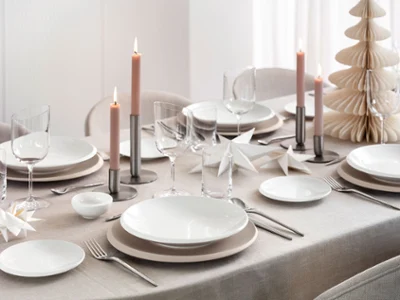 Vista cenital de una mesa festiva moderna con la colección NewMoon Beige de Villeroy & Boch: cuenco plano, platos llanos y de pan, combinados con cubertería NewMoon, copas de vino y vasos. Detalles decorativos navideños suaves, como adornos claros, ramas de pino o velas, aportan un toque estacional refinado a los tonos naturales y formas minimalistas.