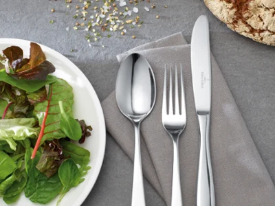 Un piatto di insalata accanto a cucchiaio, forchetta e coltello su un tovagliolo grigio su un tavolo grigio.