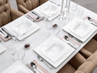 Dining table set with Modern Grace crockery A beautiful dining table setting with square white crockery from the Villeroy & Boch Modern Grace brand, silver cutlery, wine glasses and beige napkins. Beige upholstered chairs surround the table. There are three candlesticks in the centre.