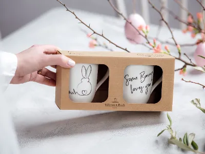 La main d’une femme tient un joli emballage cadeau avec 2 tasses ornées d’un motif de lapin. En arrière-plan, se trouvent des branches de Pâques avec des œufs roses décoratifs.