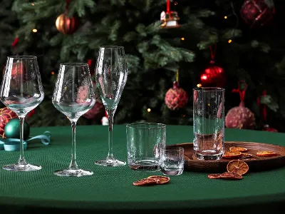 A stylish arrangement from the Villeroy & Boch Toys Delight glassware collection, consisting of wine glasses and drinking glasses, elegantly presented on a green table. Dried citrus slices are on a wooden tray in the foreground, complemented by the festive presence of a decorated Christmas tree in the background.