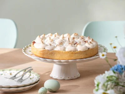 A cake with meringue stands on a wooden table on a cake stand with Easter decorations from Easter Delight, next to plates and flowers.