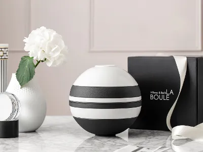 Un modèle La Boule de Villeroy & Boch en noir et blanc est posée sur une surface en marbre, elle est accompagnée d’un vase blanc avec une fleur et d’une boîte noire ouverte portant le nom « La Boule ».
