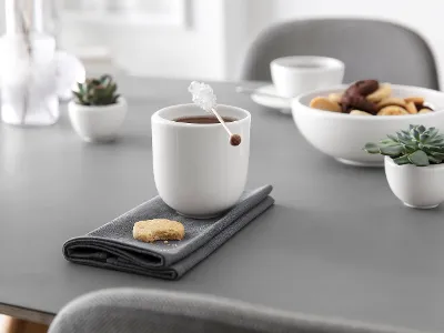 Une tasse à thé NewMoon fumante avec un bâton de cannelle, accompagnée d’un biscuit sur une serviette et de bols de biscuits et de fruits dans une cuisine calme et lumineuse.