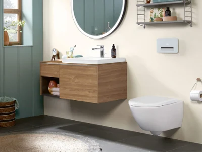 Une salle de bains moderne avec un miroir rond au-dessus d’un meuble de lavabo en bois, des toilettes suspendues et une étagère murale avec des accessoires et des serviettes. Sur le côté gauche, se trouve une fenêtre laissant entrer la lumière du soleil.