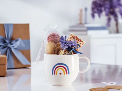 Une tasse « With Love » de Villeroy & Boch au design arc-en-ciel, remplie de macarons, d’une tablette de chocolat et avec fleurs violettes sur une surface blanche avec des boîtes cadeaux et des livres en arrière-plan.