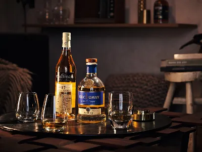 Two bottles of Scotch whisky and three Villeroy & Boch glasses on a wooden table. Books and a decorative figurine are visible in the background.