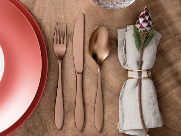 A place setting with a red plate, rose gold fork, knife, and spoon, a glass, and a napkin with a floral napkin ring on a wooden table.
