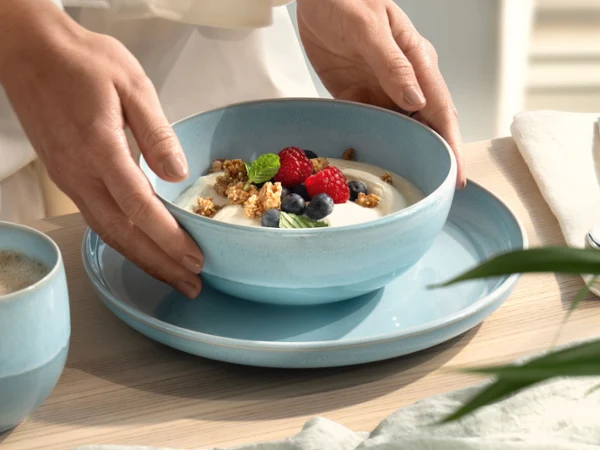 Hände, die eine blaue Perlemor Aqua Schale mit Joghurt, Müsli, Blaubeeren und Himbeeren auf einen blauen Teller stellen.
