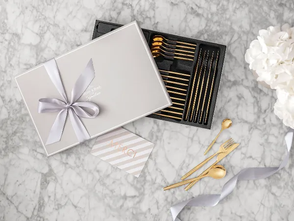 Gift box with gold MetroChic cutlery and a bow, a thank you card and white flowers on a marble surface.