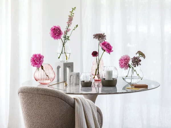 A marble table with various flowers in Rose Garden glass vases and Rose Garden hurricane lamps in front of a window with curtains.