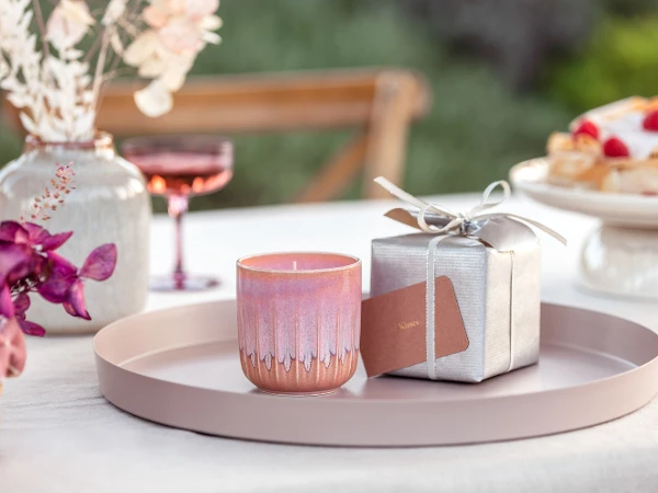 Scented candle from the like. by Villeroy & Boch collection as a gift on a round tray with a silver gift box in the background and pink orchids in the foreground.