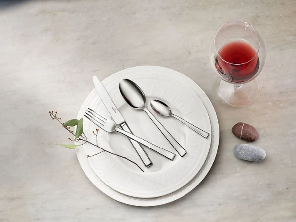 Ein Besteckset von Victor mit Messer, Gabel und zwei Löffeln liegt auf weißen Cellini Tellern, neben einem Glas Rotwein und zwei glatten Steinen auf einer Marmoroberfläche.