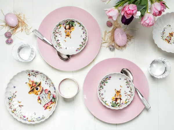 Deux couverts dressés avec des assiettes roses, des plats creux décorés de motifs de lapin, des cuillères, des verres à eau, une bougie et des œufs décoratifs de la collection Spring Fantasy de Villeroy & Boch. En haut à droite, on peut voir un bouquet de tulipes roses.