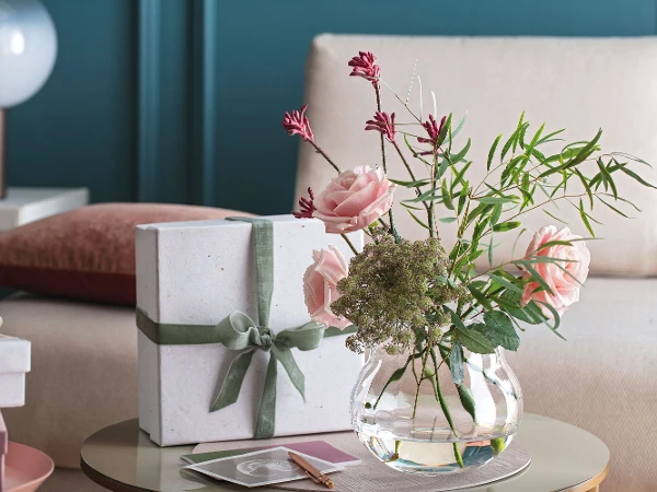 Un vase Rose Garden avec des fleurs roses et un cadeau sur une table.