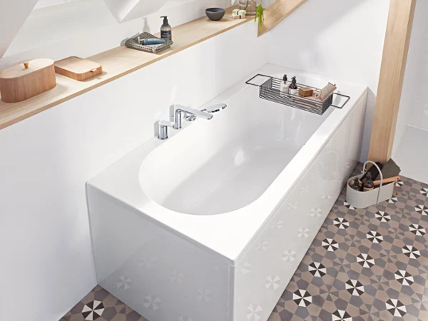 A white bath from the O.novo collection with a silver tap standing next to a wooden shelf with toiletries. The bathroom has patterned tiles and natural light comes in from a window above the bath.