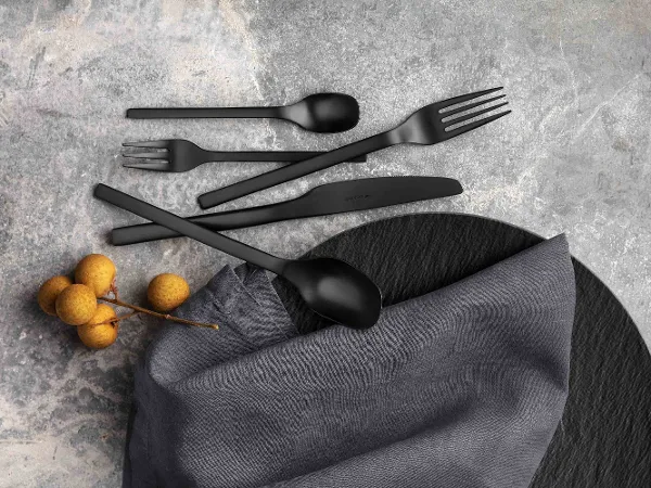 Black Manufacture flatware set with fabric napkin and decorative berries on a textured gray surface.