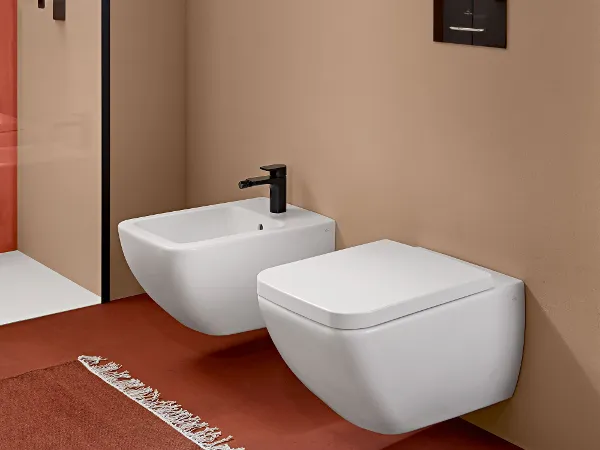 Une salle de bains moderne avec un bidet et des WC fixés au mur, une étagère avec des serviettes pliées et une cabine de douche en verre. La pièce a des murs bruns et un sol rouge avec un tapis à motifs.