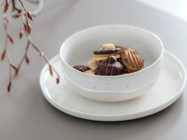 A Winter Glow serving bowl with various cookies on a Winter Glow plate with decorative branches in the background.