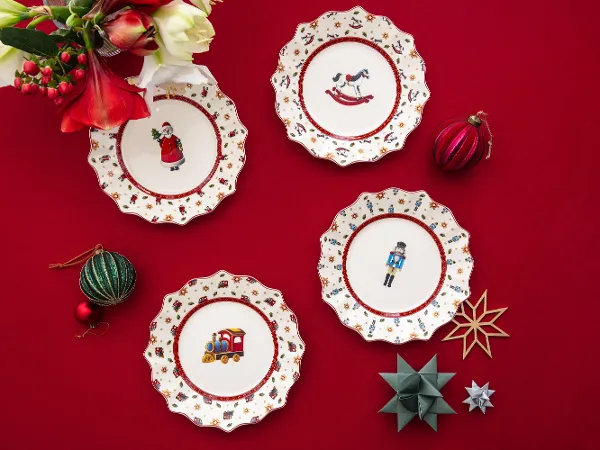 Four Christmas plates from the Toys Delight anniversary collection on a red tablecloth Four decorative Christmas plates with festive motifs on a red tablecloth, surrounded by ornaments and flowers.