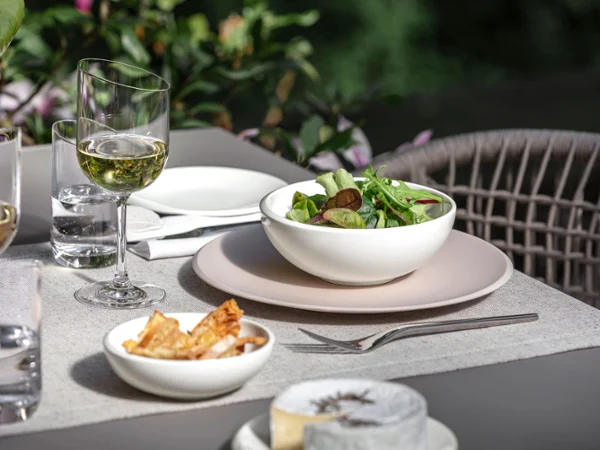 White NewMoon bowl with salad presented on a beige dinner plate A Villeroy & Boch NewMoon bowl filled with salad beside wine glasses.