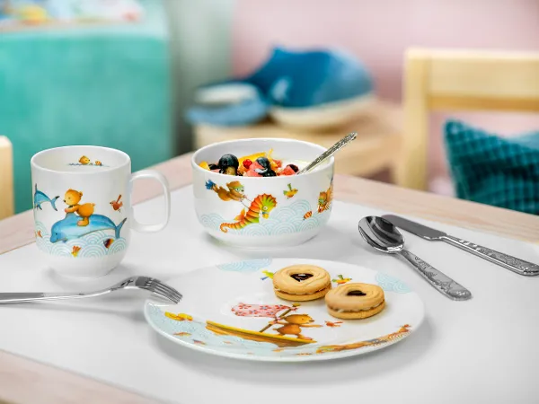 Une présentation de petit-déjeuner avec une salade de fruits, des biscuits sur une assiette et deux tasses de vaisselle pour enfants Happy as a bear sur une table.