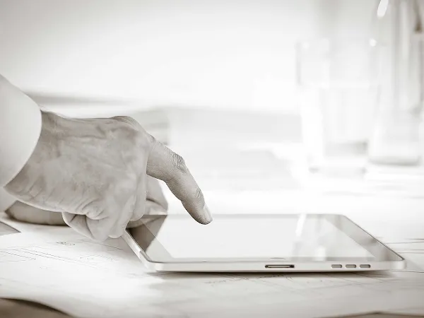 Eine Nahaufnahme einer Person, die mit ihrem Zeigefinger den Bildschirm eines Tablets berührt. Im Hintergrund sind Papiere und ein Glas Wasser zu sehen.