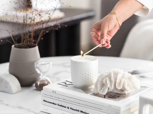 Une personne allume une bougie parfumée Manufacture de couleur blanche sur une table basse avec des objets décoratifs et des livres.