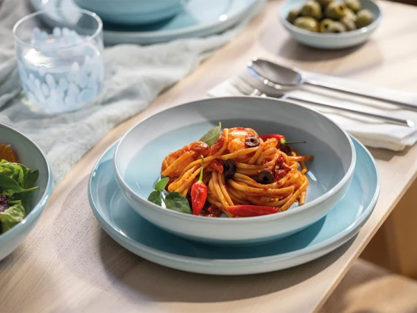 Una ciotola di pasta della collezione Perlemor Aqua con spaghetti, salsa di pomodoro e verdure è posizionata su un piatto blu Perlemor Aqua, accanto a posate e un'insalata.