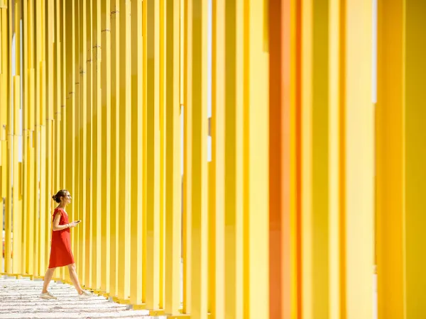 A person in a red dress walks along a row of tall, yellow columns on a sunny day.