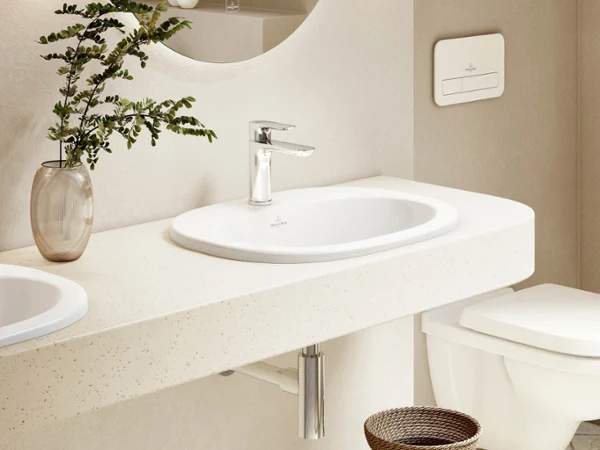 A bathroom with a white O.novo washbasin, a round mirror, a tap and a wall-mounted toilet. There is a potted plant on the shelf and a wicker basket on the floor.