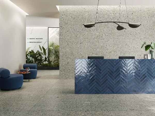 Modern hotel lobby with a structured blue reception desk, minimalist furniture, hanging lamps and a corner full of green plants.