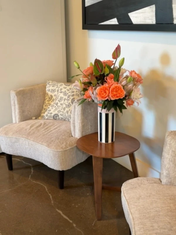 Memphis Vase with arranged flowers Two beige chairs with a patterned pillow sit by a small wooden table holding a vase of pink and orange flowers, against a light wall and polished floor.