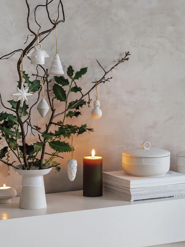 Moderne, minimalistische kerstdecoratie op een witte plank met Winter Glow vaas, takken en hangende ornamenten. 