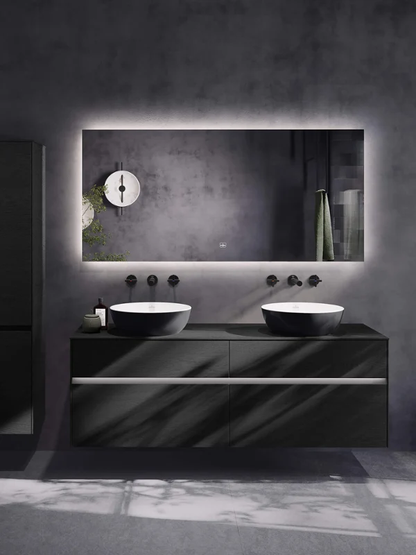 Baño moderno con lavabo doble, espejo grande retroiluminado, armarios negros y grifos minimalistas frente a paredes grises.