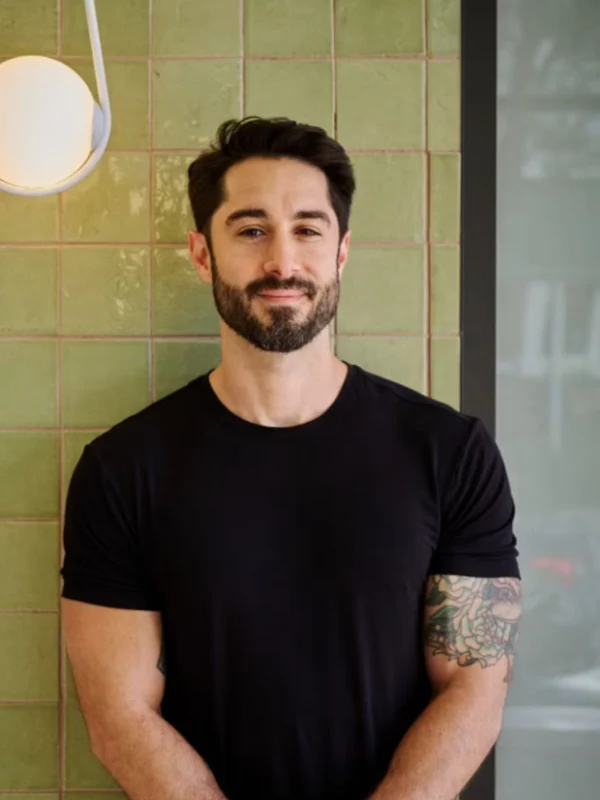 Chef Joe Anthony of Arvine Restaurant Smiling bearded man in a black T-shirt stands with folded hands before a green tiled wall, showing a tattooed arm; calm, friendly tone.